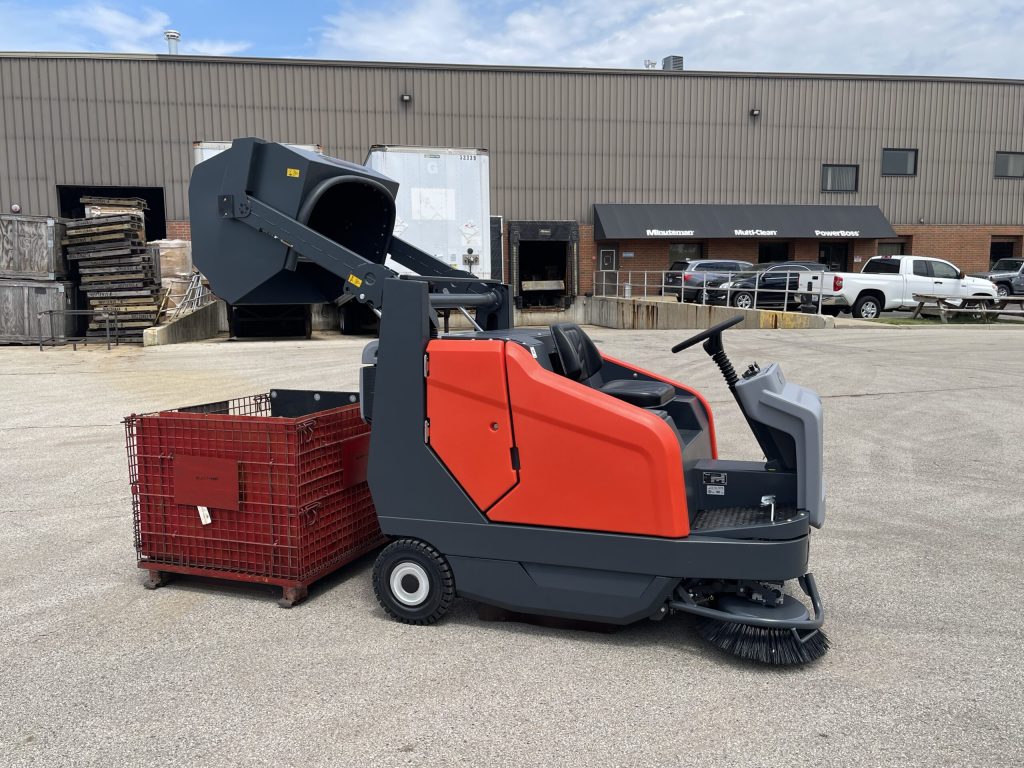 A red and gray Sweepmaster ride-on floor sweeper empties debris into a metal bin outside an industrial building on a paved lot.
